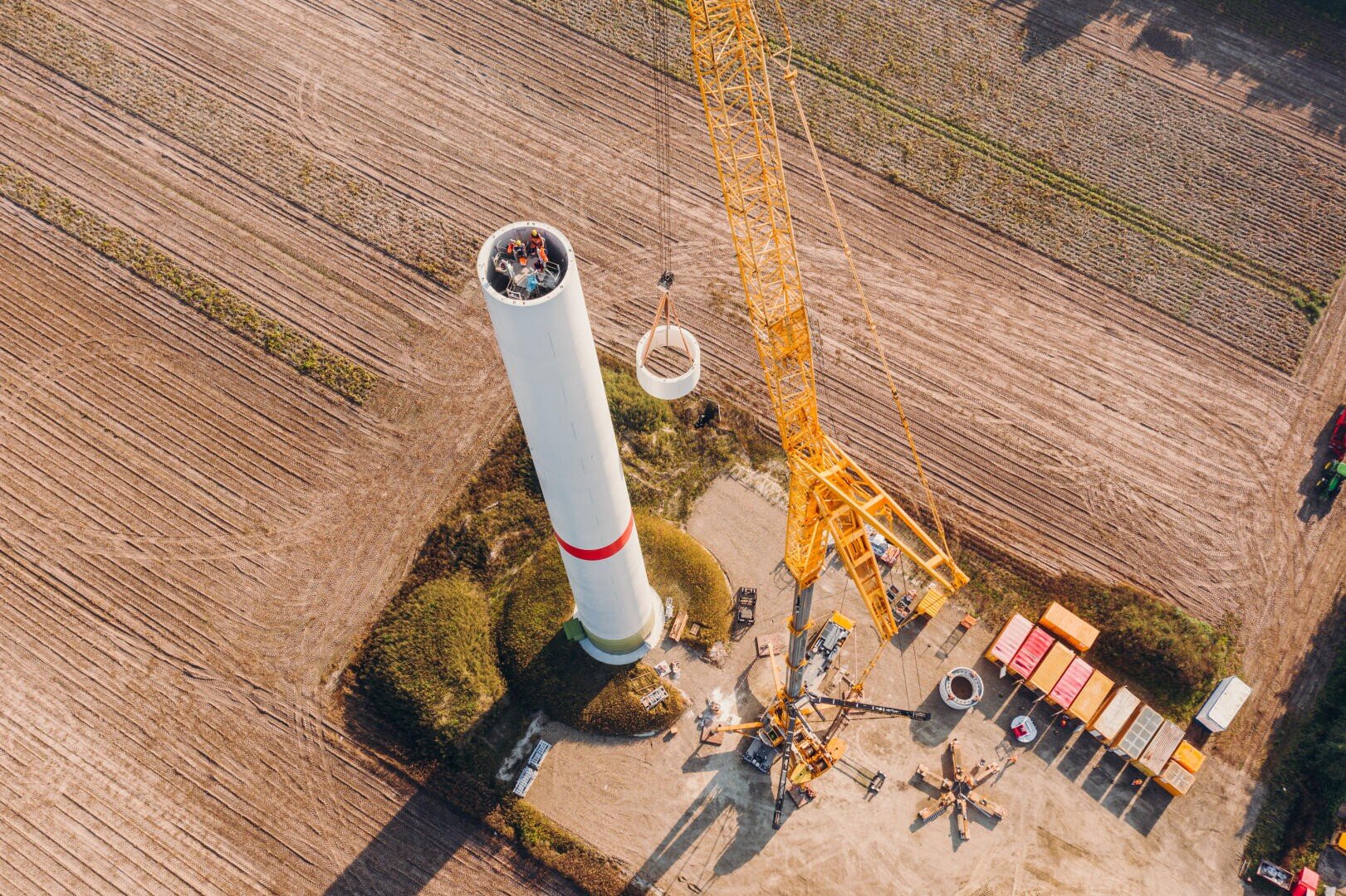 Aerial view of workers assembling a wind turbine tower in a field using a large yellow crane, surrounded by containers, equipment, and dirt roads.