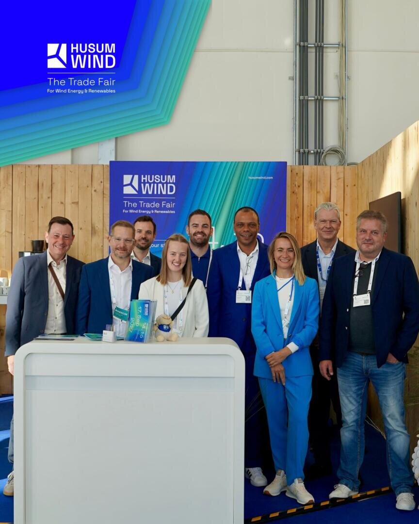 Acht Personen – sechs Männer und zwei Frauen – stehen lächelnd an einem Messestand, hinter ihnen ist ein „Husum Wind“-Banner zu sehen. Die Gruppe trägt Geschäftskleidung, und auf der Theke des Standes liegen Werbematerialien aus.