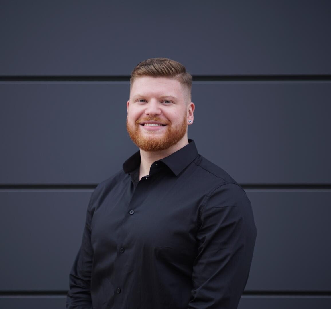 A man with short red hair and a beard, wearing a black button-up shirt, stands smiling in front of a dark gray paneled wall.