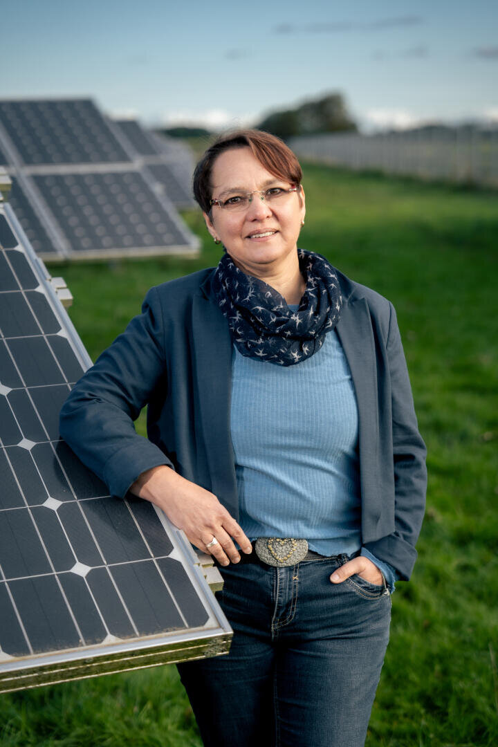 A woman wearing glasses, a blue blazer and a scarf is standing outside, leaning on a solar panel. She is smiling, and in the background you can see a meadow and more solar panels.