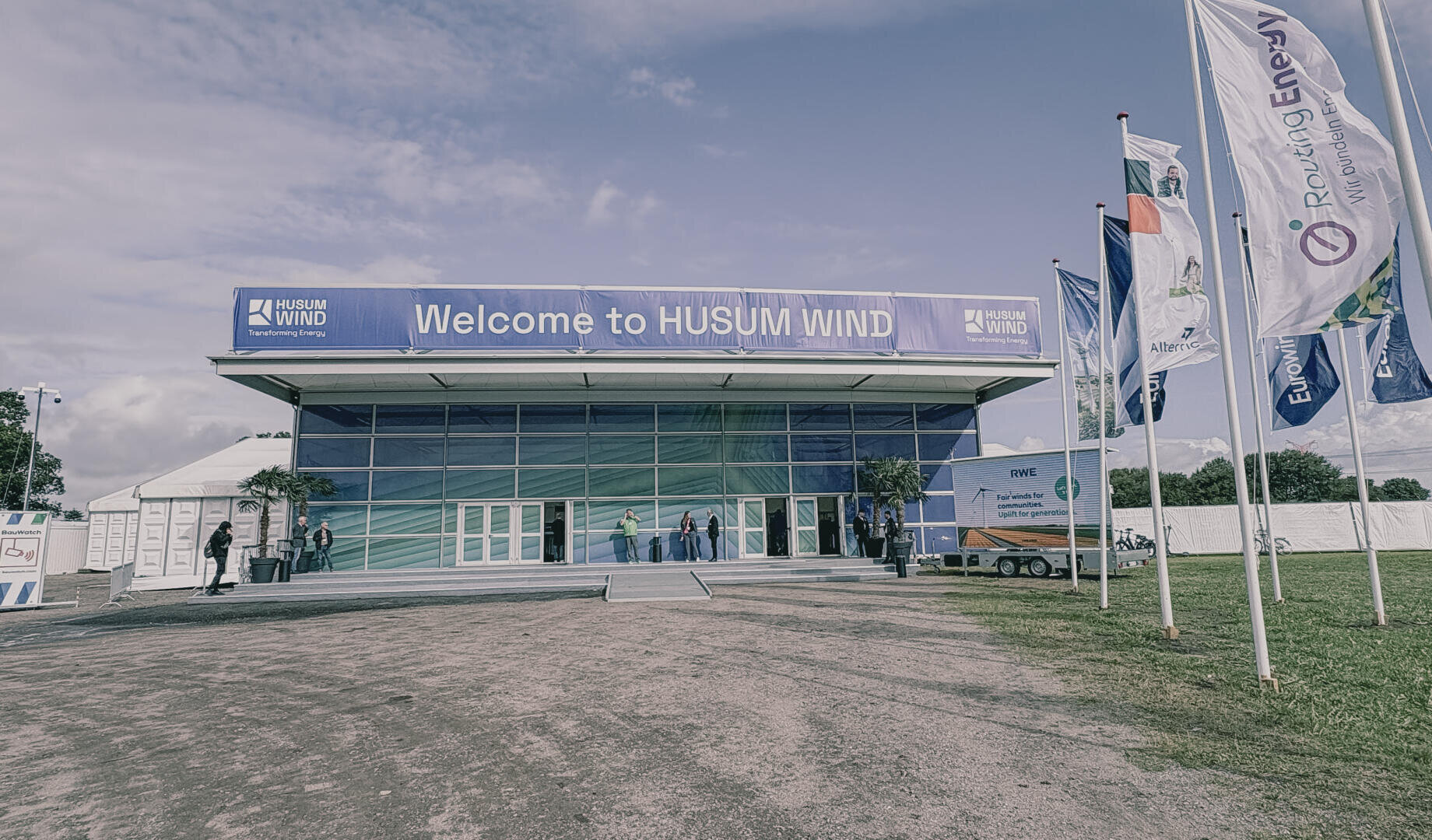 A large building with a sign saying Welcome to HUSUM WIND at the entrance. Several people are standing near the doors and there are large flags with various logos on the right-hand side. The sky is partly cloudy.