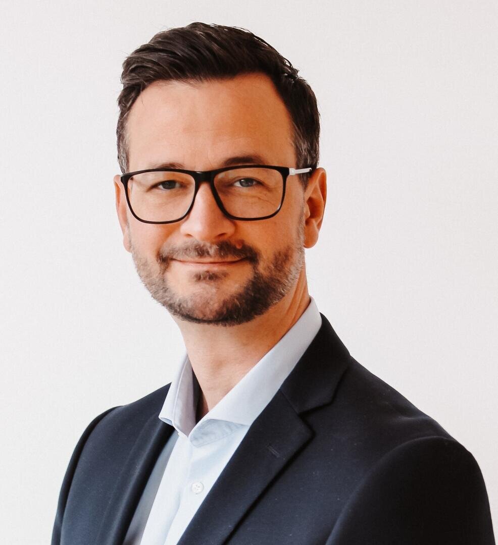 A man with short dark hair, a beard, and glasses is wearing a dark suit jacket over a light blue shirt, smiling slightly, and posing against a plain white background.