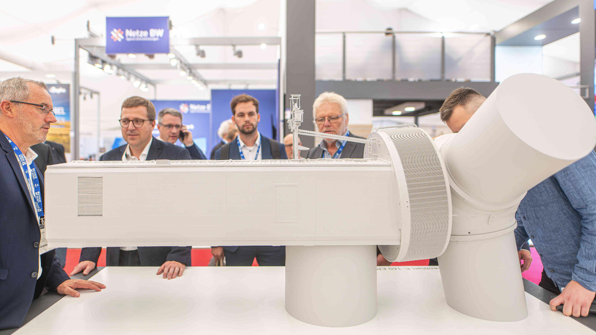 Mehrere Männer in Businesskleidung betrachten auf einer Indoor-Ausstellung oder Messe ein großes, detailliertes Modell einer Windkraftanlagengondel. Im Hintergrund sind Messestände und Banner zu sehen.