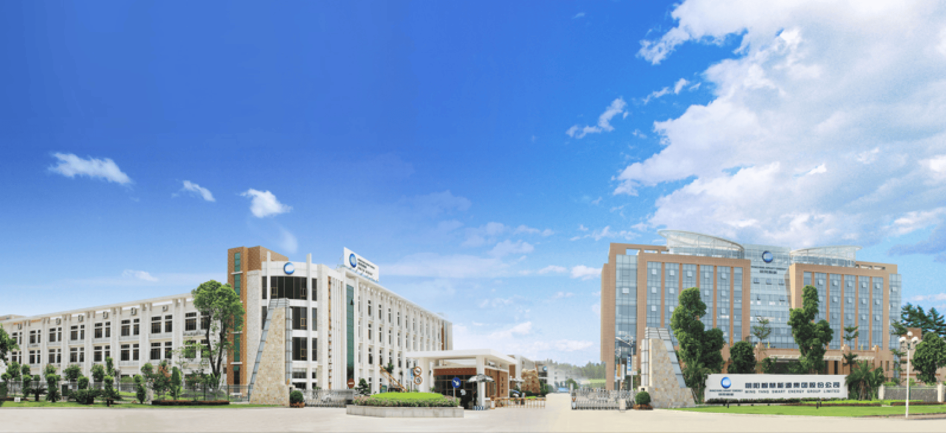 Wide view of a modern campus with two large multi-storey buildings, green spaces and a blue sky with scattered clouds. Signs and flags can be seen at the entrance.