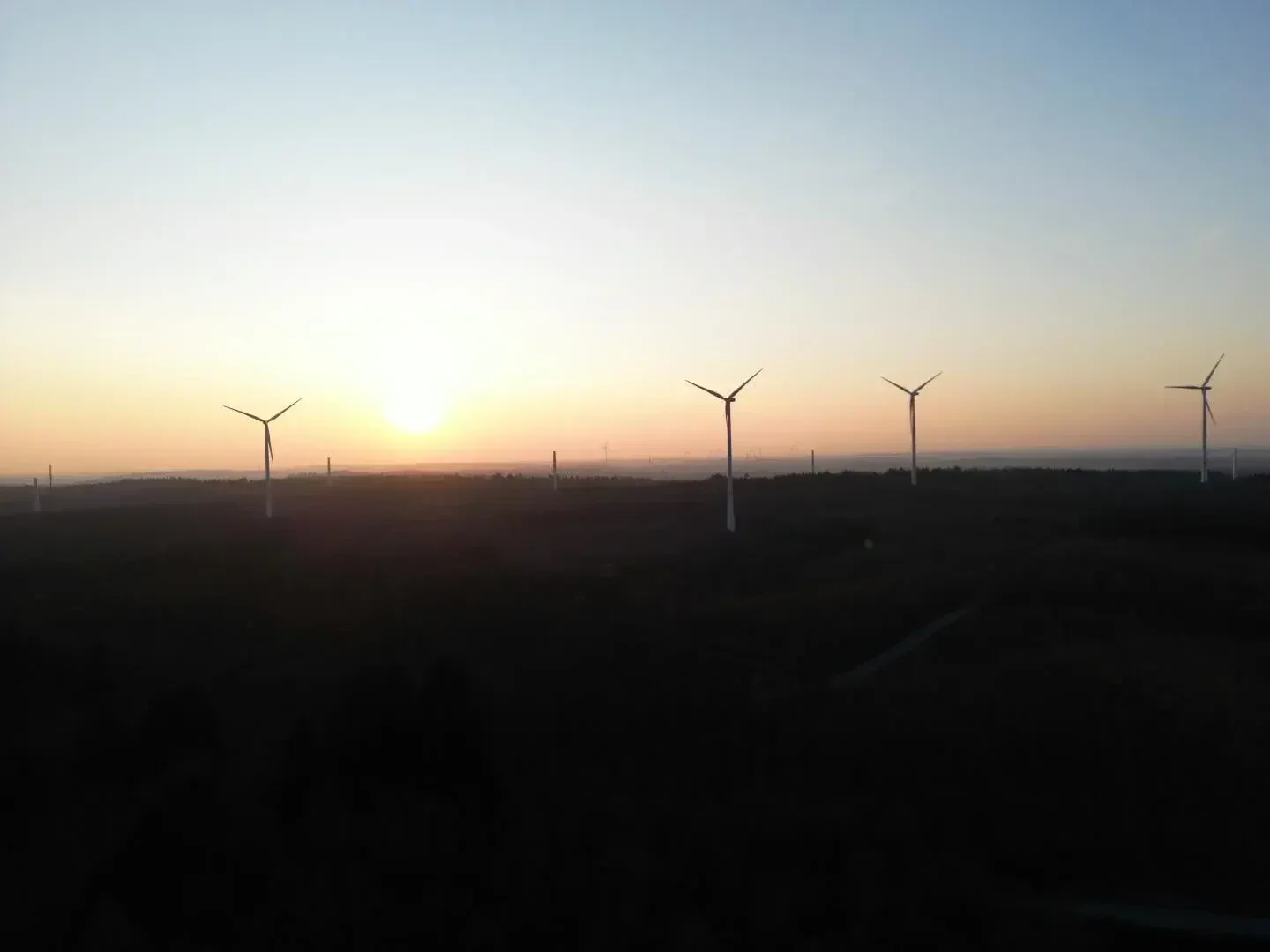 Die Windturbinen heben sich bei Sonnenuntergang als Silhouette vom Himmel ab und stehen in einer dunklen Landschaft, während die Sonne tief am Horizont steht und ein sanftes orangefarbenes Licht wirft.