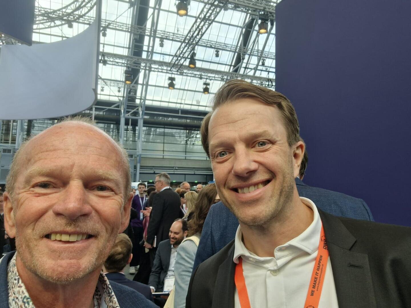 Zwei lächelnde Männer machen ein Selfie bei einer belebten Indoor-Veranstaltung für Windkraftbranche mit Menschen im Hintergrund. Der Veranstaltungsort hat eine Glasdecke und ein modernes Metallgerüst. Ein Mann trägt ein geblümtes Hemd, der andere ein weißes Hemd und ein orangefarbenes Schlüsselband.