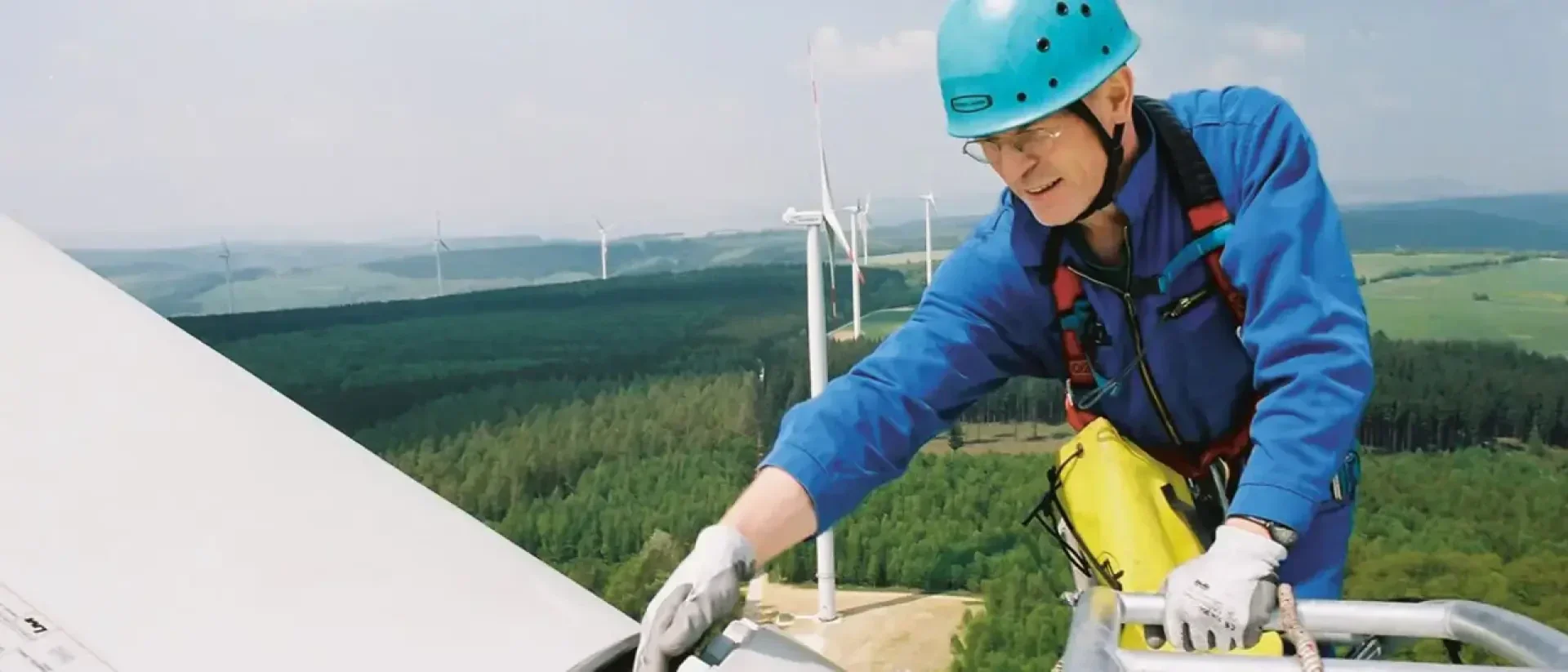 A worker in blue protective clothing and helmet inspects a wind turbine blade on a high platform, while several wind turbines and a wooded landscape can be seen in the background.