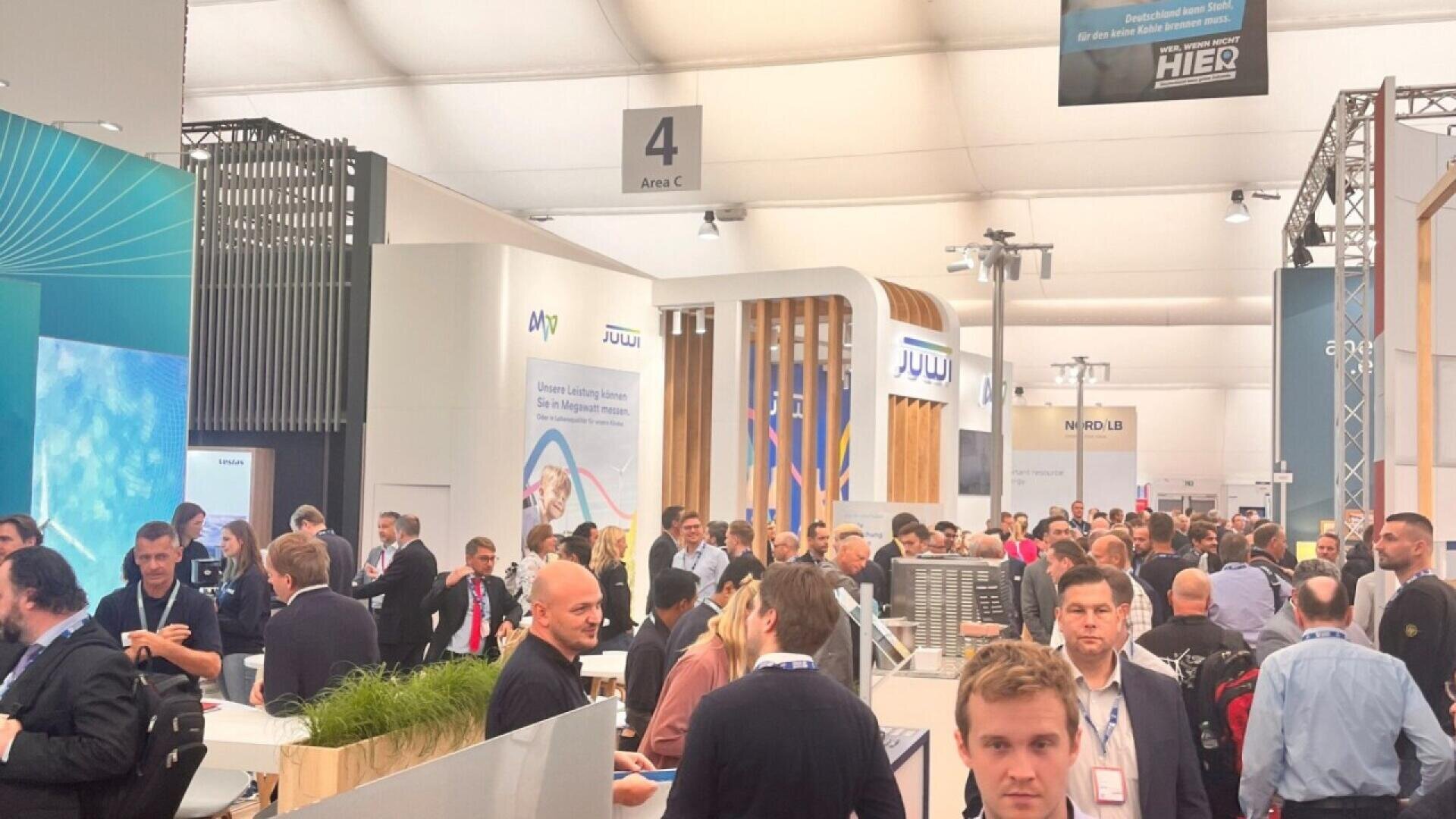 A crowded exhibition hall filled with people walking and talking, surrounded by booths and displays under a large white tent. Signs and banners with company logos and a 4 section marker are visible overhead.