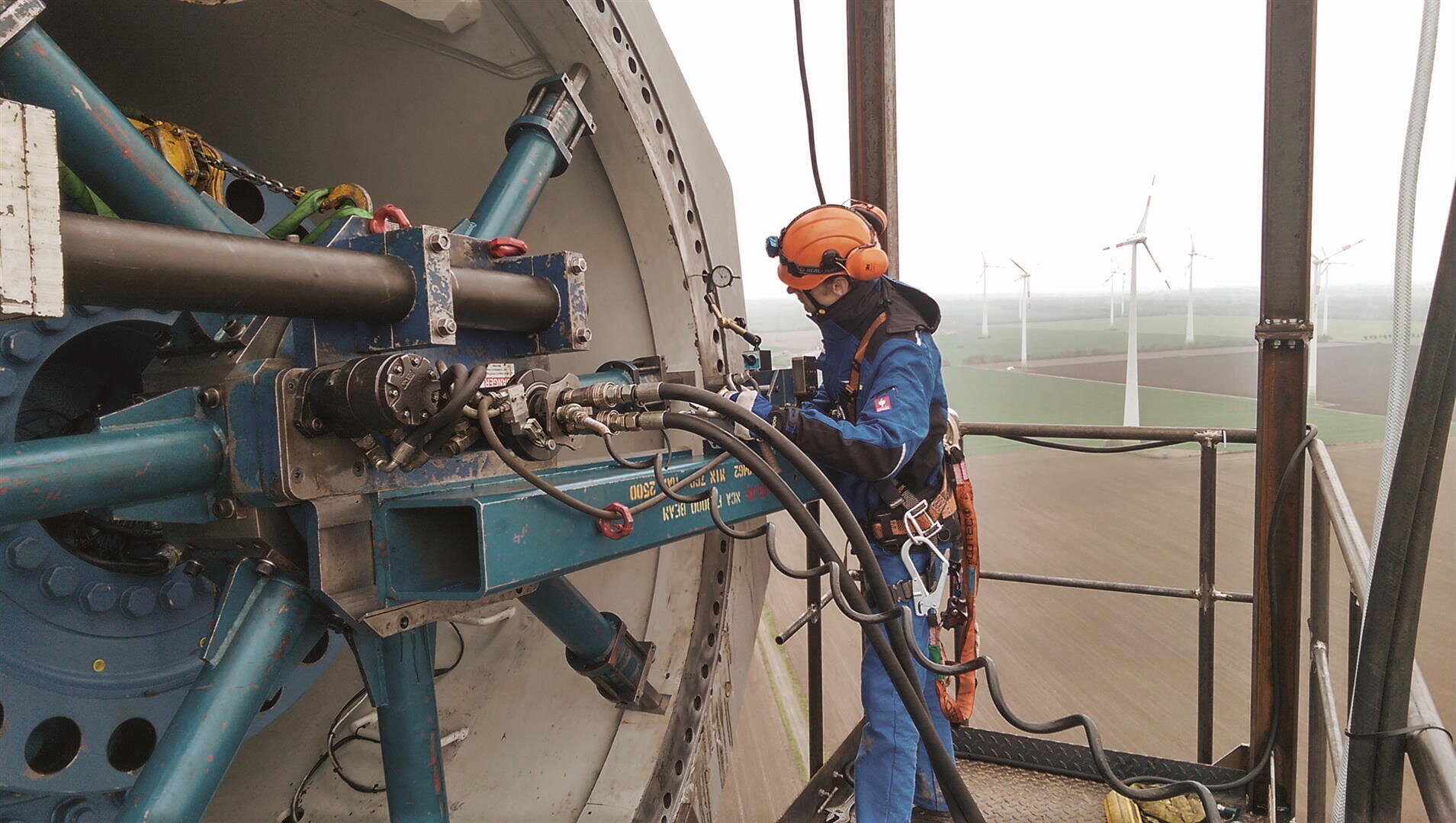 Ein Techniker in Schutzkleidung arbeitet an den Maschinen auf der Spitze einer Windkraftanlage, während im Hintergrund mehrere Windkraftanlagen in einer flachen Landschaft zu sehen sind.