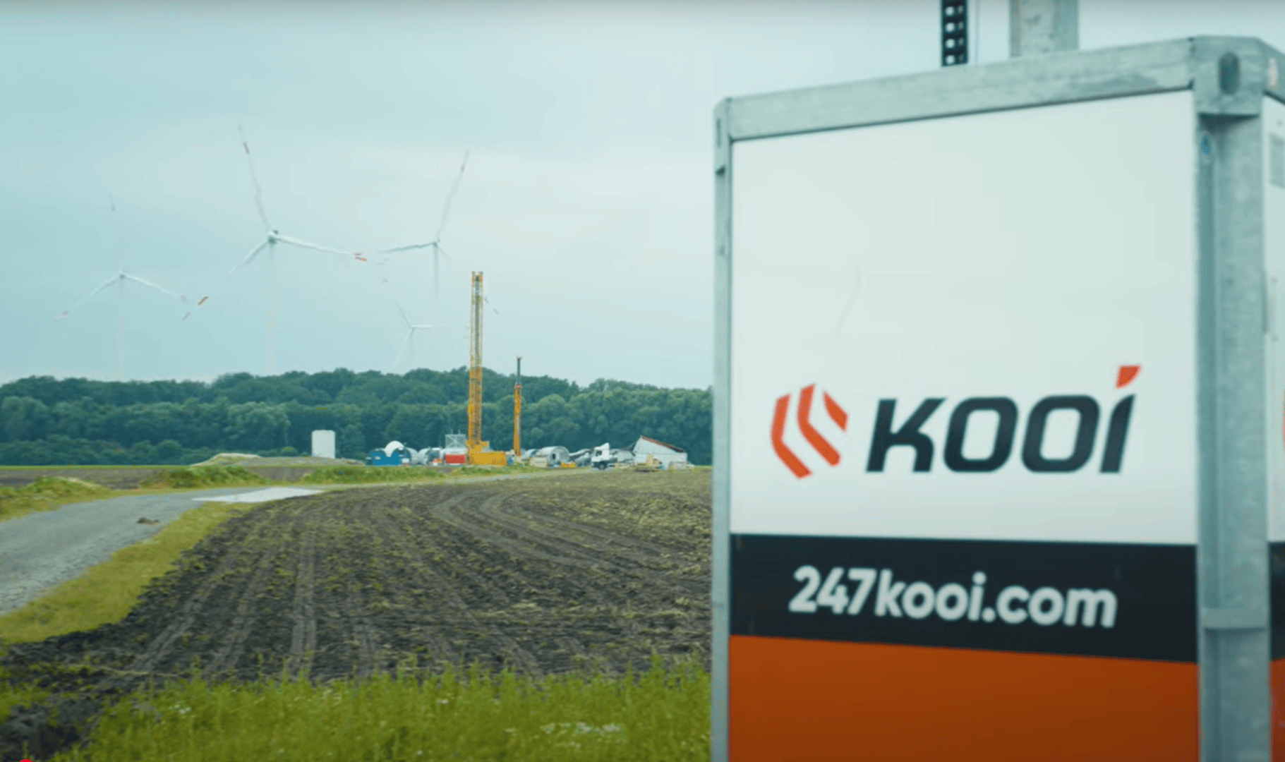 Ein Schild mit dem Logo Kooi und der Website 247kooi.com steht im Vordergrund einer Baustelle mit Windrädern, Kränen und Bäumen im Hintergrund unter einem bewölkten Himmel.