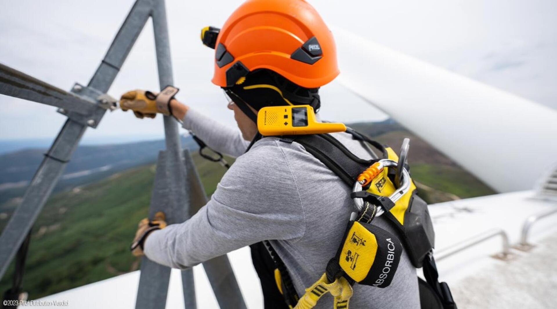 Ein Arbeiter, der einen Helm und eine Sicherheitsausrüstung trägt, benutzt einen Gurt, während er an einer Metallkonstruktion hoch über dem Boden arbeitet, und hat ein gelbes Walkie-Talkie an seiner Weste befestigt.