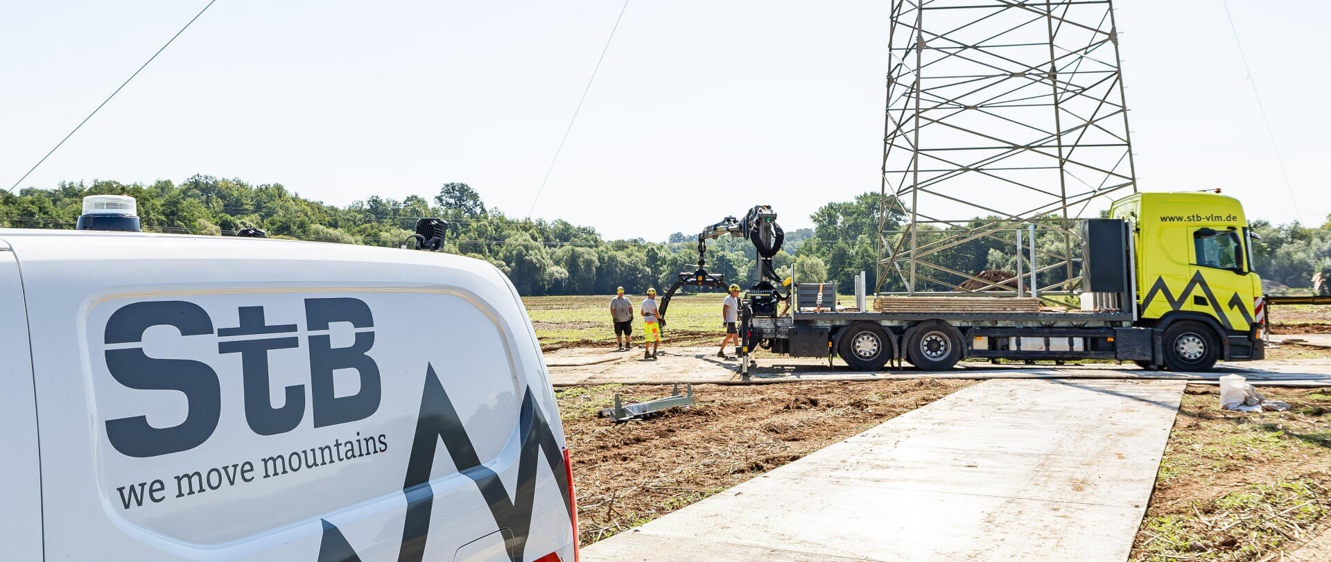 A white van with StB wir versetzen Berge is parked near a construction site where workers and a yellow truck with a crane are standing by a large metal tower in an open field.