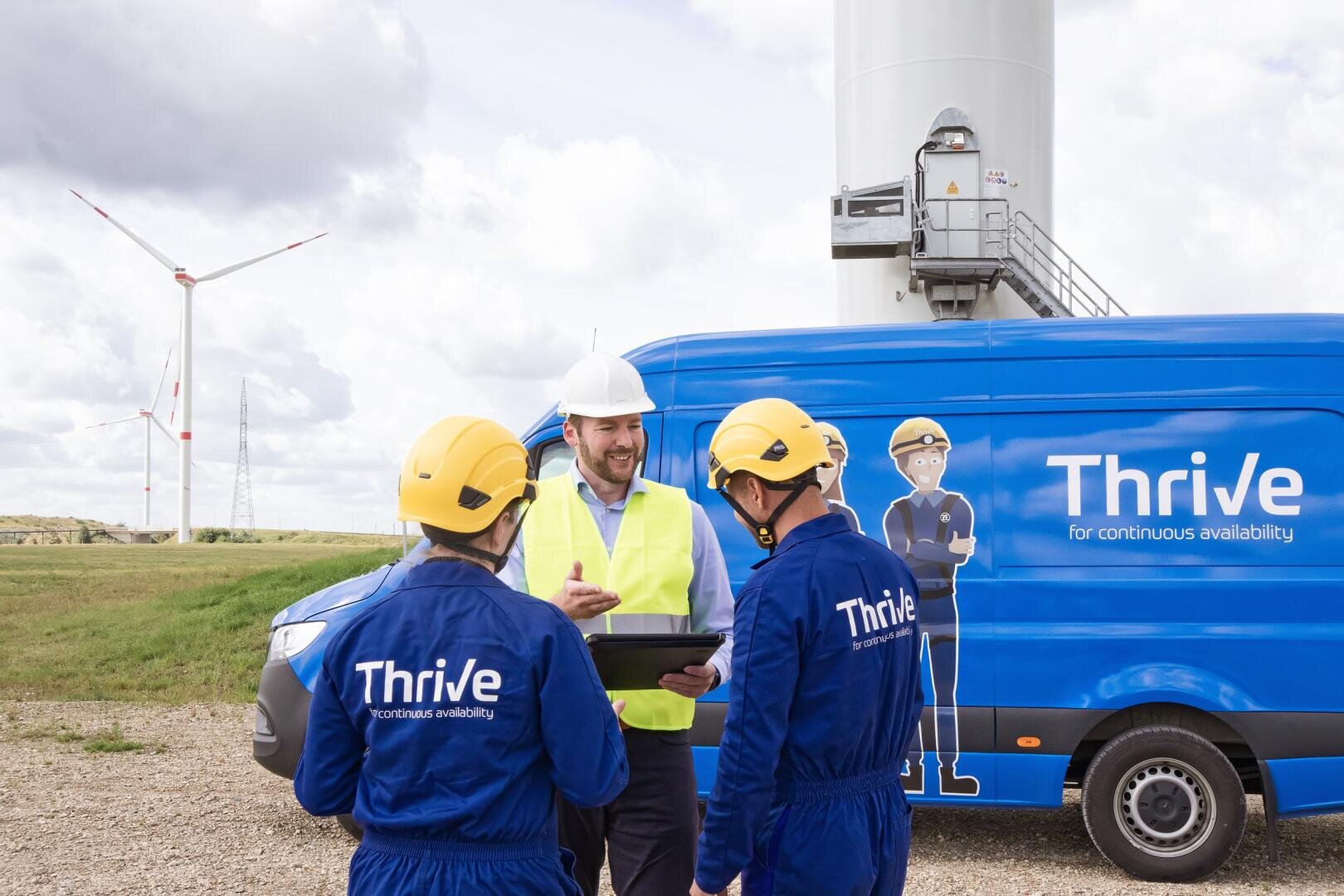 Three workers in safety gear, two in blue Thrive uniforms and one in a yellow vest and white helmet, stand beside a blue Thrive van and a wind turbine, discussing something on a tablet. Wind turbines are visible in the background.