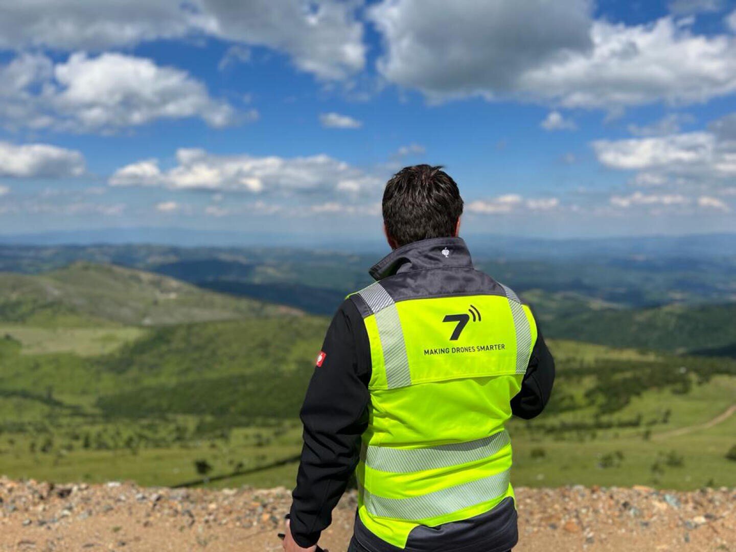 A person in a high-visibility vest stands on a hilltop, looking out over a vast, green landscape under a blue sky with scattered clouds. The vest reads 7 MAKING DRINKS SMARTER on the back.