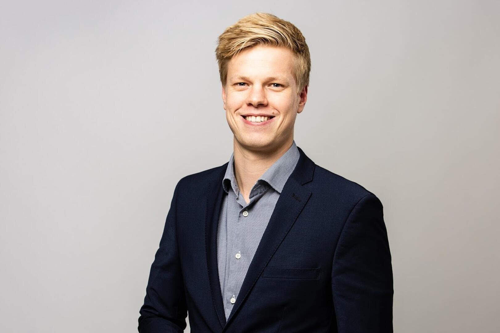 A young man with blond hair wearing a dark suit jacket and light blue dress shirt smiles confidently at the camera against a plain light gray background.