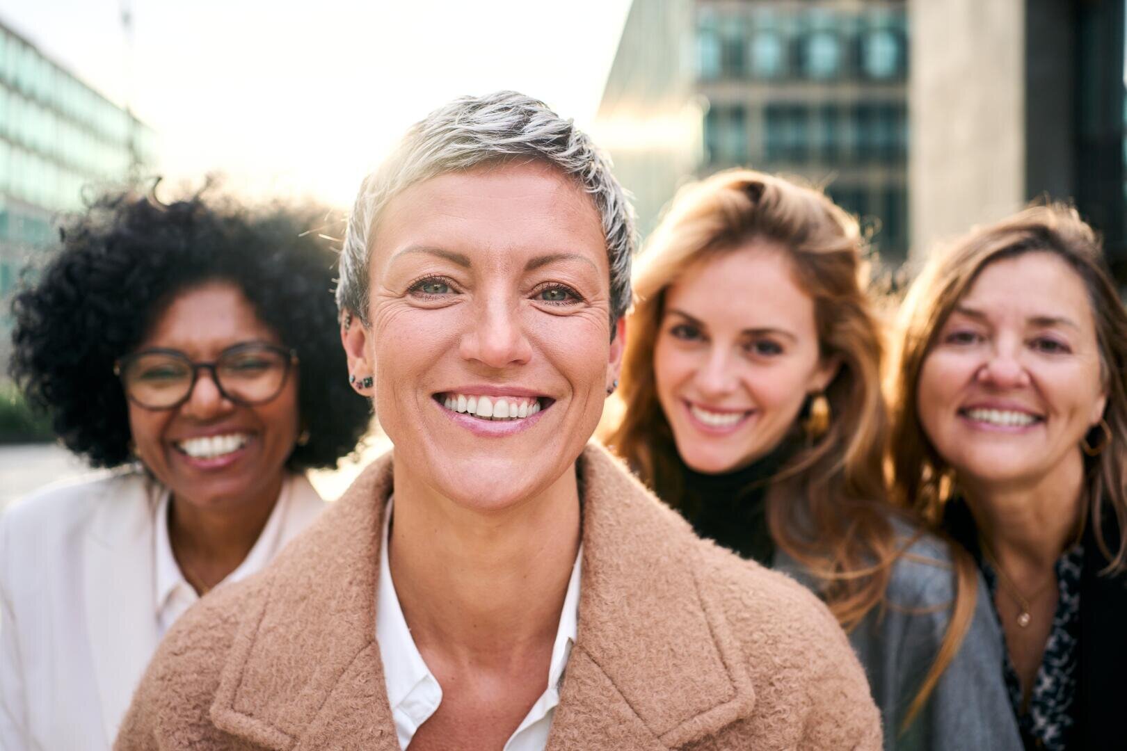 Four women stand close together outdoors and smile at the camera. The sunlight shines behind them and creates a warm and cheerful atmosphere. They appear cheerful and friendly, dressed in casual and business attire.