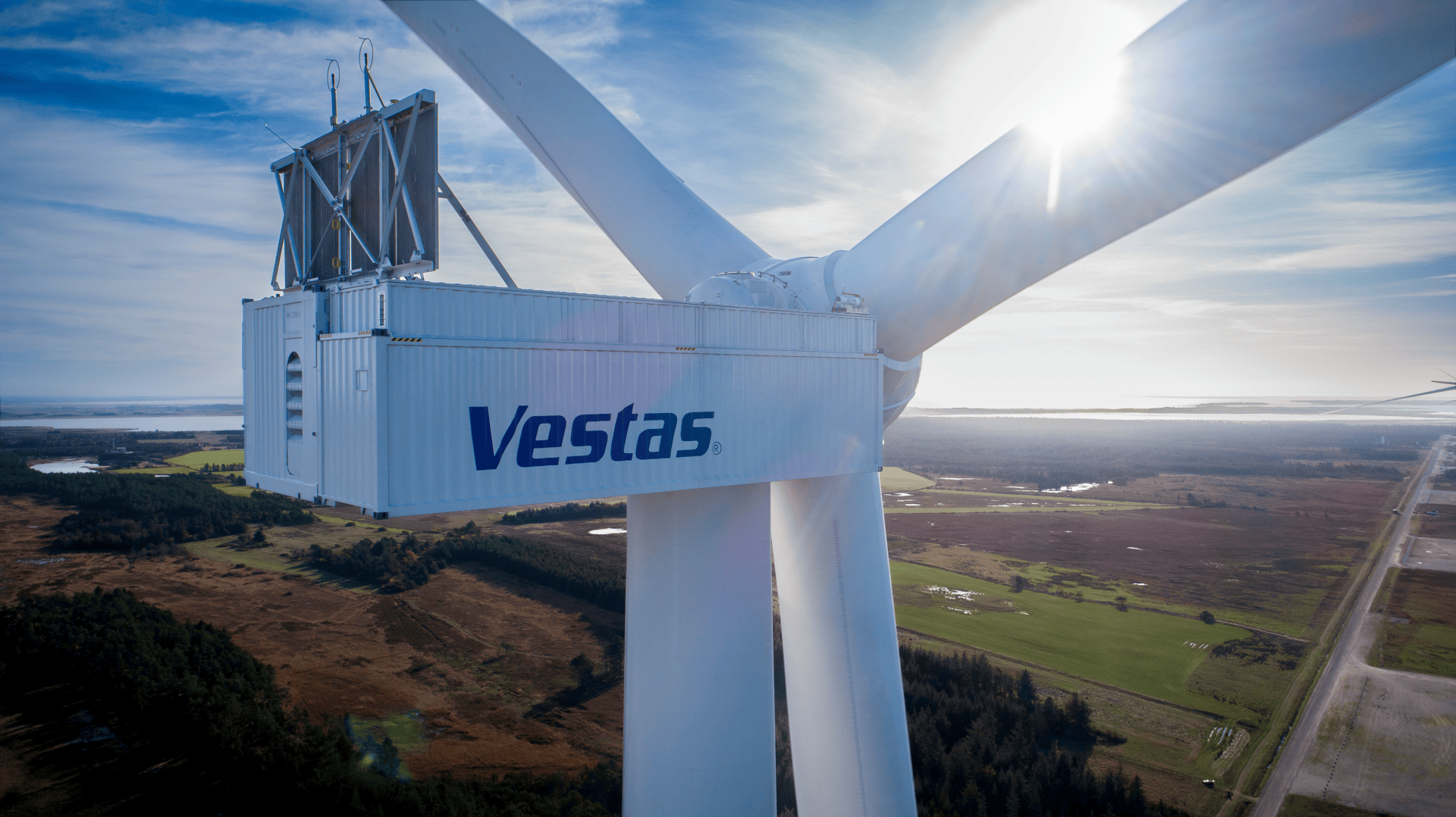 Eine Nahaufnahme einer großen weißen Vestas-Windturbine vor einem sonnigen Himmel mit Blick auf weite Felder, Wälder und Wasser in der Ferne.