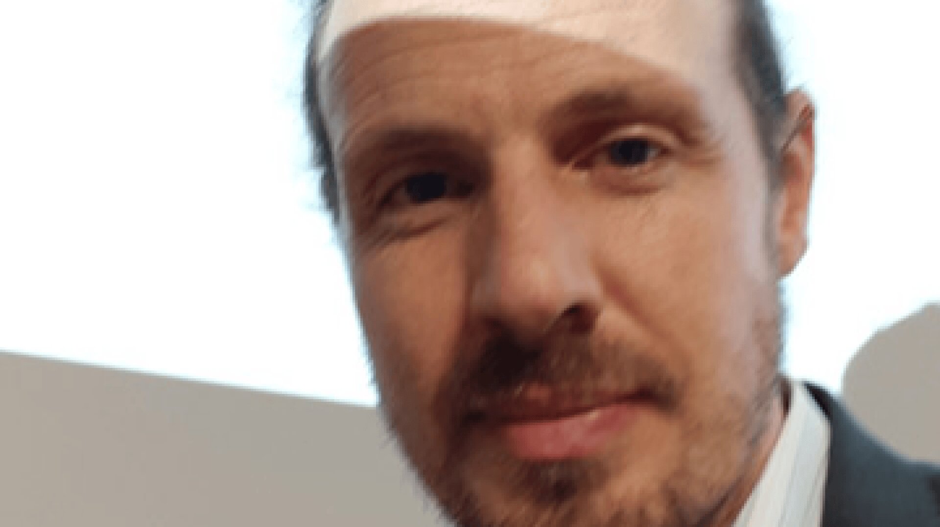 A close-up of a man with light skin, a short beard, and mustache, wearing a collared shirt and jacket, with a neutral expression and a bright background.