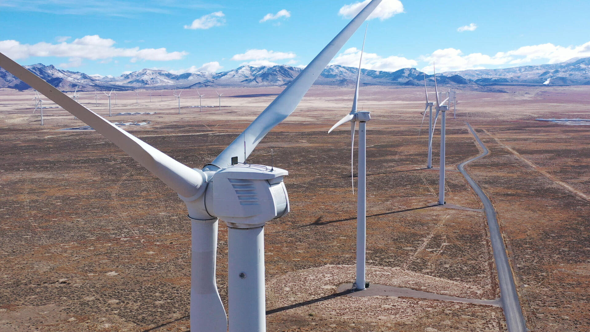 Close-up of wind turbines in a dry, open plain with a road and snow-capped mountains in the background under a partly cloudy blue sky.