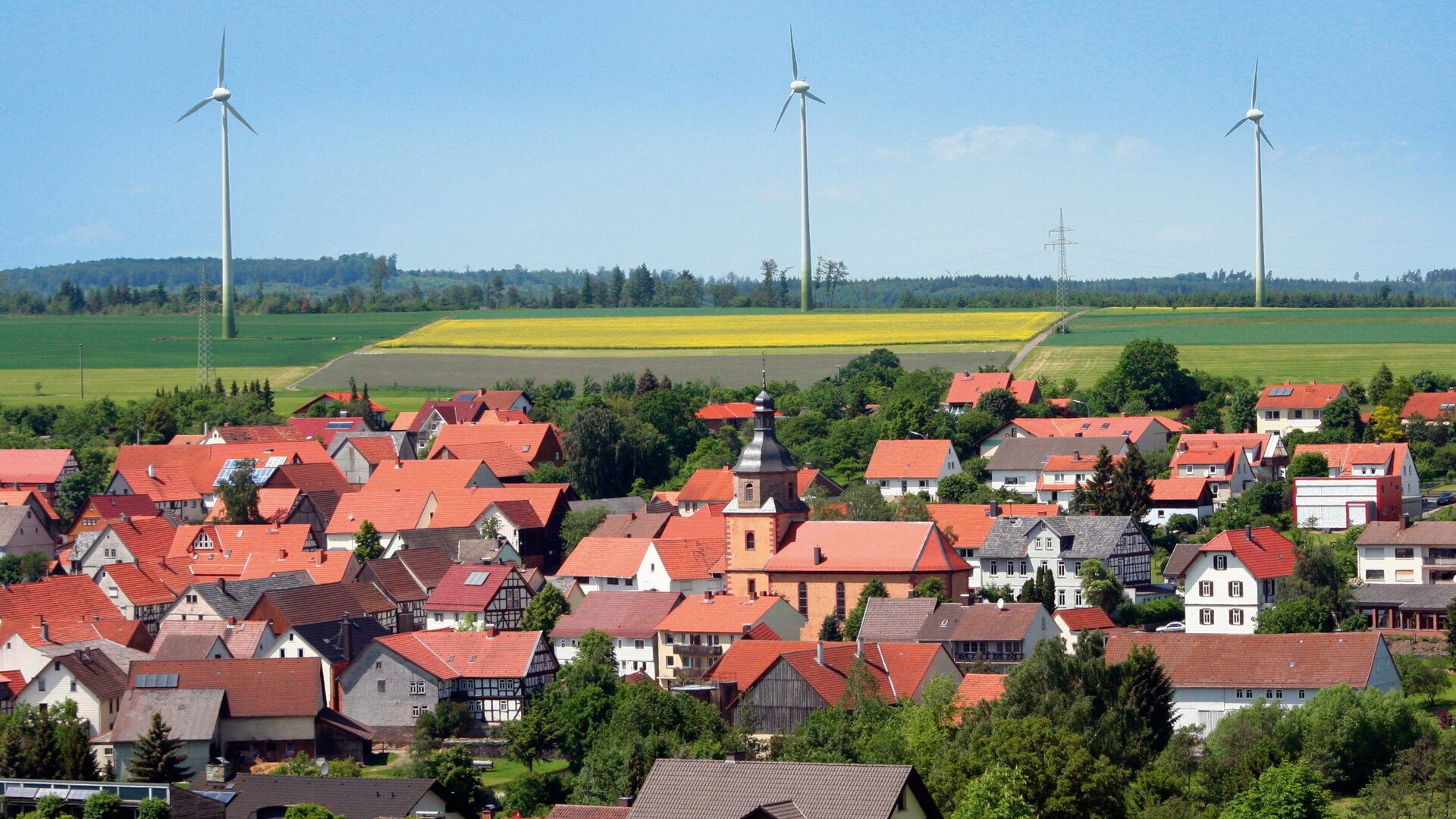Ein Dorf mit roten Häusern und einer Kirche, umgeben von grünen Feldern und Bäumen, mit drei Windturbinen im Hintergrund unter einem blauen Himmel.