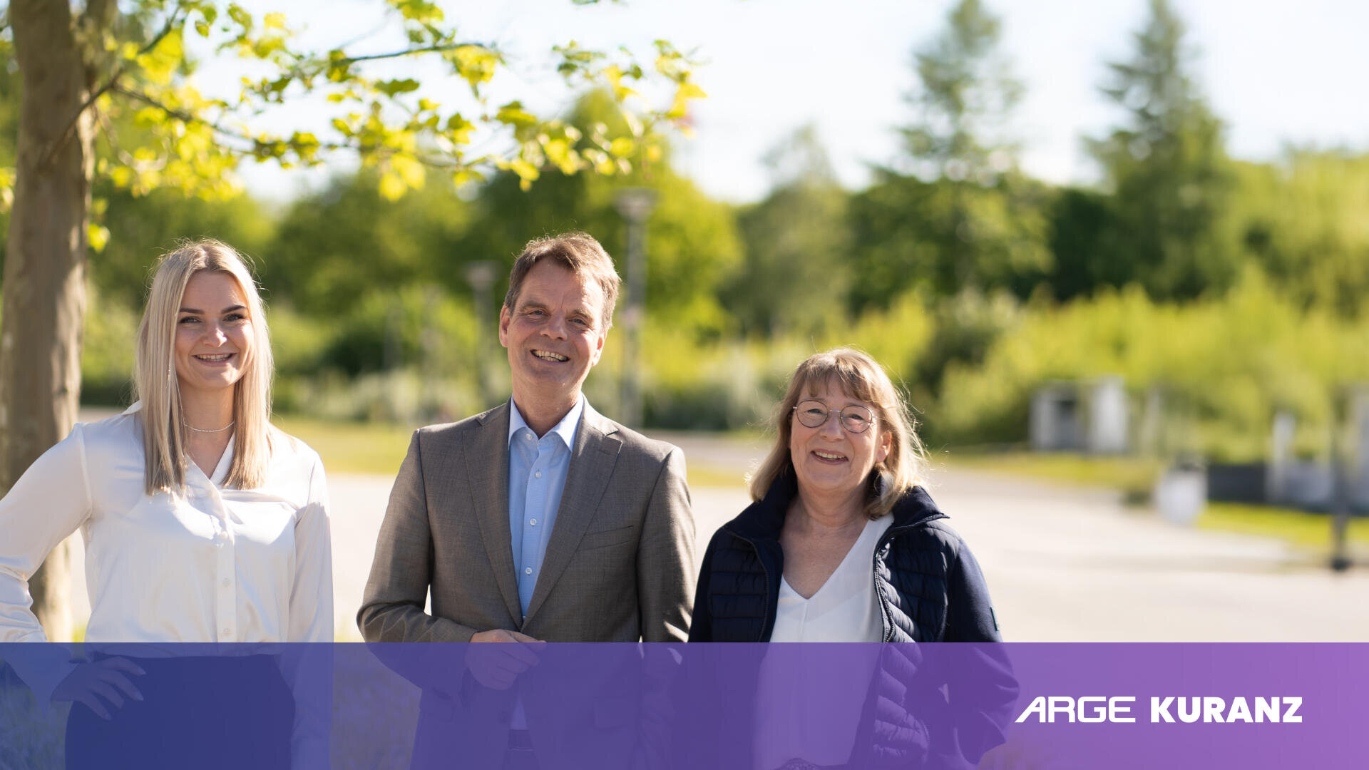 Drei Personen - zwei Frauen und ein Mann - stehen lächelnd im Freien, mit Bäumen und Grünpflanzen im Hintergrund. Ein violettes Banner mit der Aufschrift ARGE KURANZ erscheint unten rechts im Bild.
