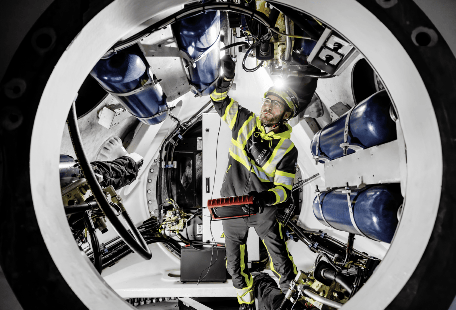 A worker in protective clothing inspects blue tanks and equipment in a large, circular industrial machine illuminated by skylights.