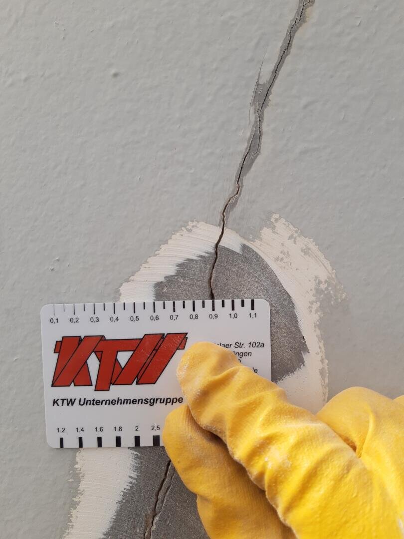 Eine behandschuhte Hand hält eine Messkarte gegen einen Mauerriss, auf der die Breite des Risses neben einer geflickten Stelle angegeben ist. Die Karte ist mit einem Lineal und dem Logo der KTW Unternehmensgruppe versehen.