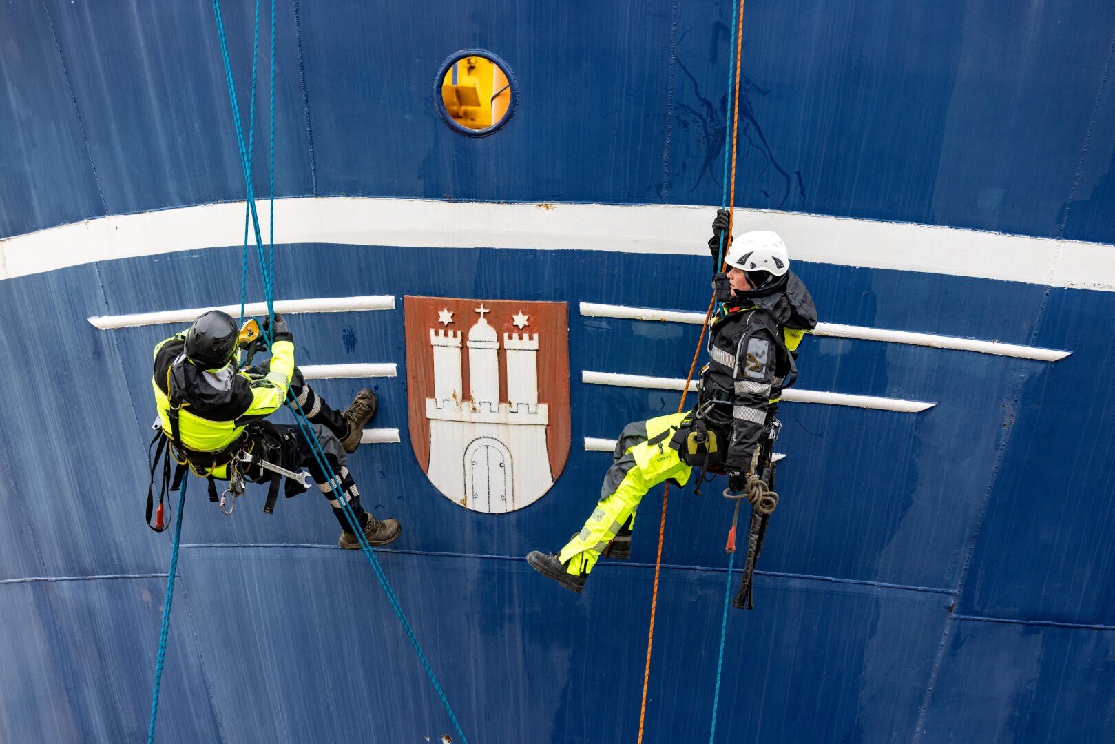 Two workers in protective gear are suspended from ropes as they work on the side of a large blue ship with a white and red coat of arms showing a castle and two towers.
