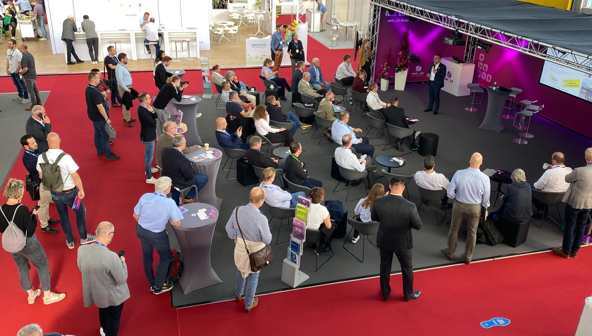 Eine Gruppe von Menschen sitzt und steht bei einer Indoor-Veranstaltung um eine kleine Bühne herum und hört einem Redner vor einem lila Hintergrund mit Bildschirmen und Lichtern zu. Die meisten Teilnehmer stehen mit dem Gesicht zur Bühne auf einem roten Teppichboden.