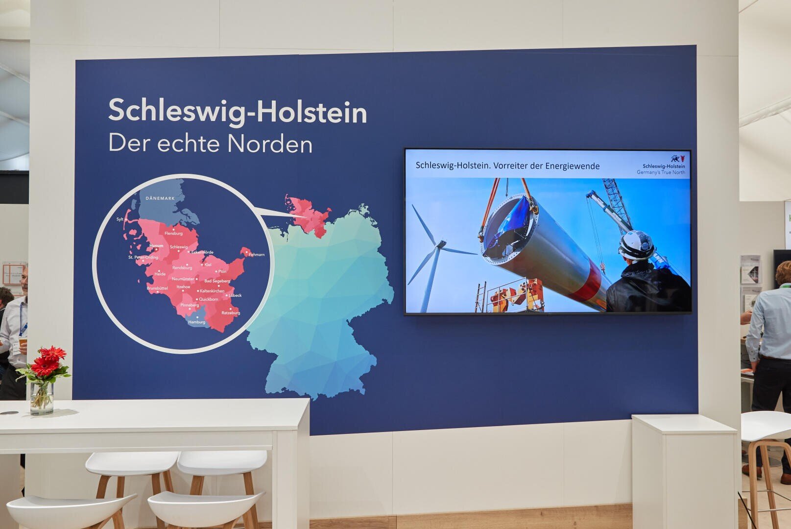 An exhibition stand shows a map of Germany on which Schleswig-Holstein is highlighted in red, as well as a TV screen with a picture of wind turbines and workers. The text reads: Schleswig-Holstein. The real north.