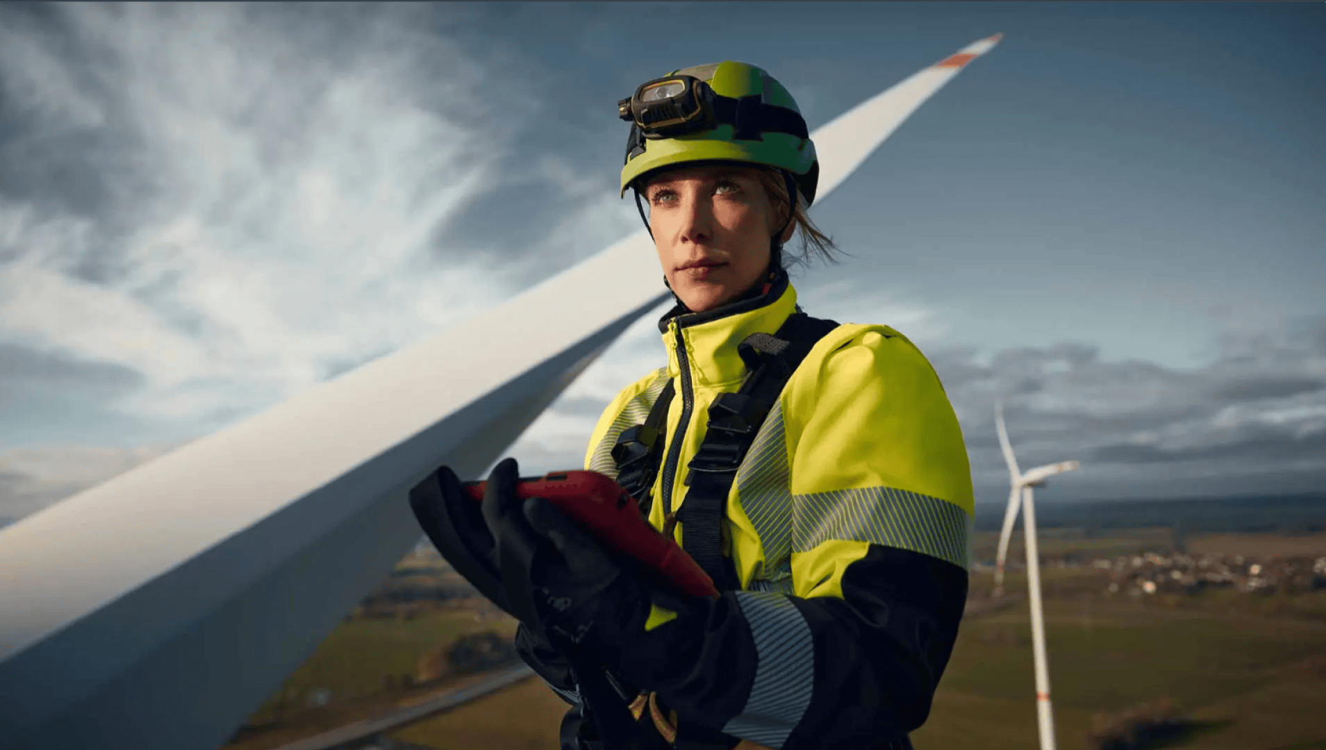 A worker wearing a high-visibility suit, helmet and gloves stands near a wind turbine, holding a tablet and looking ahead. The sky is partly cloudy and other wind turbines can be seen in the background.