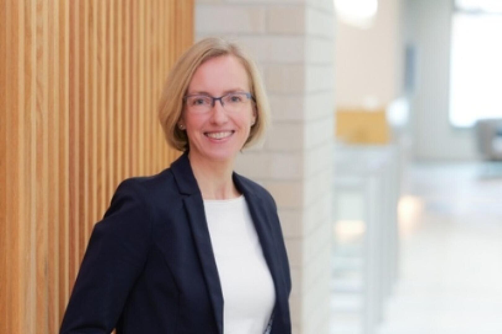 Eine Frau mit kurzen blonden Haaren und Brille, die einen dunklen Blazer über einem weißen Oberteil trägt, steht in einem hell erleuchteten, modernen Raum an einer Holzwand und lächelt in die Kamera.