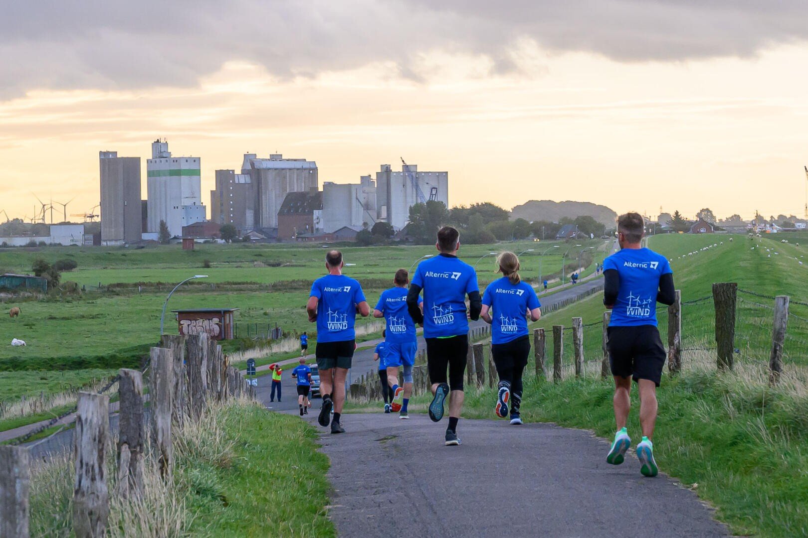 A group of runners in blue shirts jog along a country lane lined with grass and fences, with industrial buildings and a cloudy sky in the background.