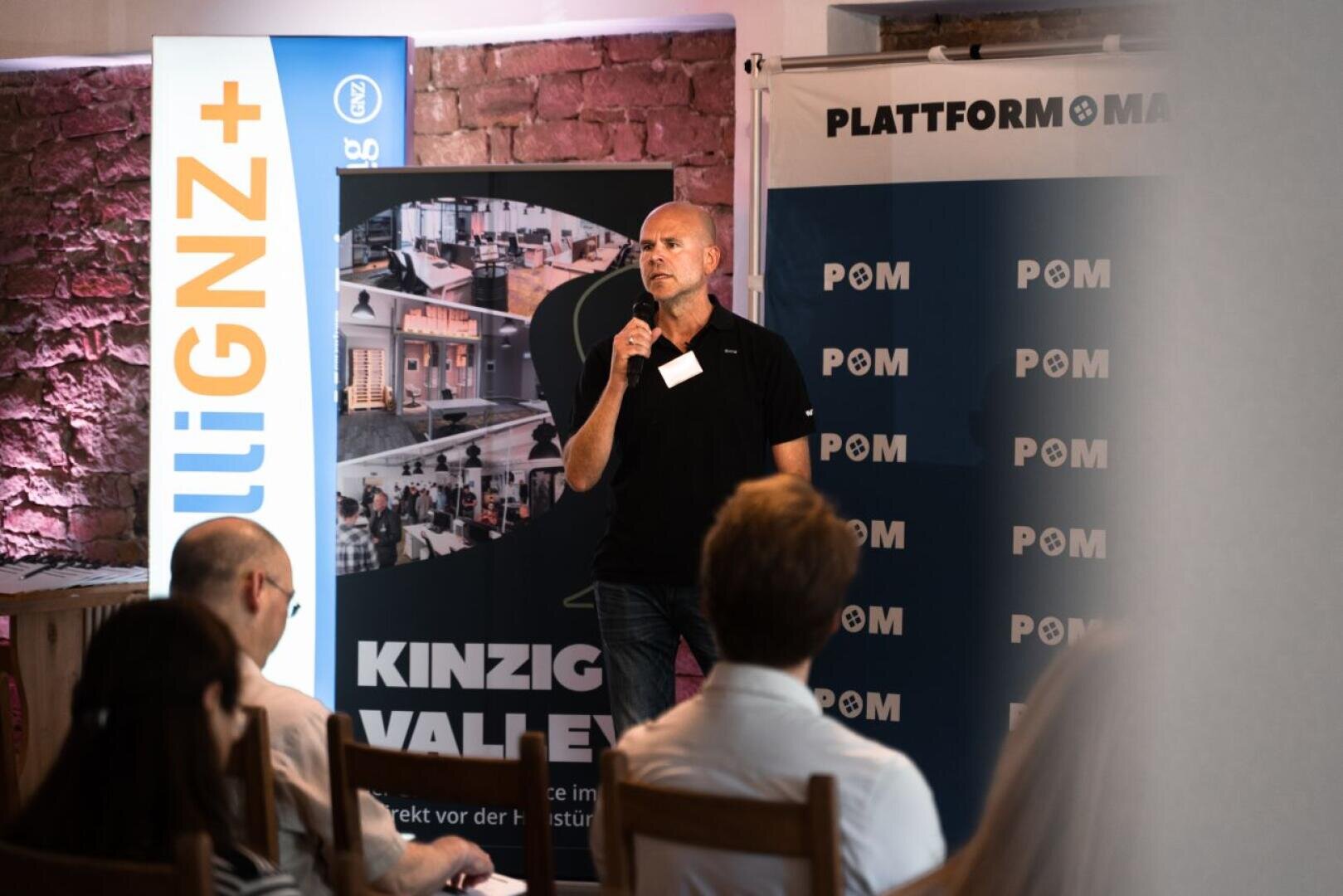 A man with a microphone speaks to the audience at an indoor event, who are standing in front of banners with the words PLATTFORM and KINZIG VALLEY. Several people sit opposite him and listen attentively.