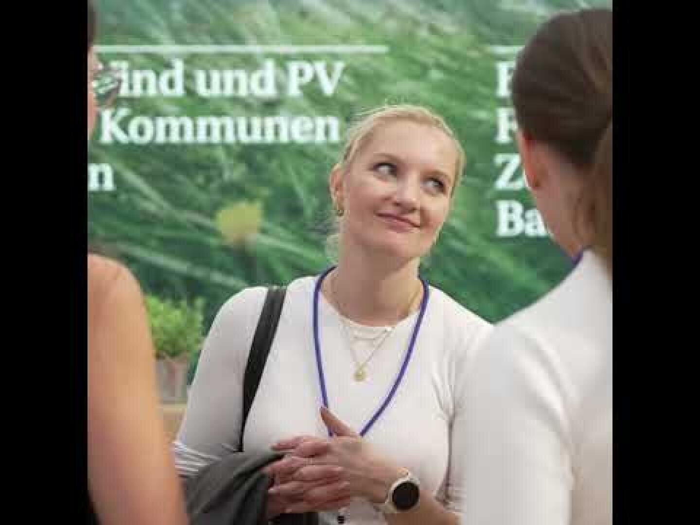 Eine Frau mit zurückgebundenen blonden Haaren, die ein weißes Oberteil und ein blaues Schlüsselband trägt, lächelt im Gespräch mit zwei anderen Personen. Grüner Text ist im unscharfen Hintergrund teilweise sichtbar.