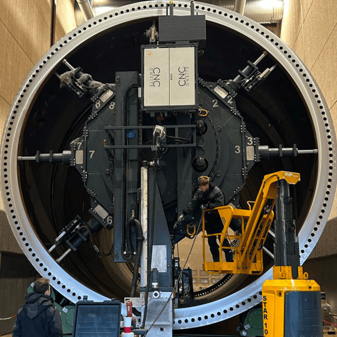 Ein Arbeiter steht auf einem gelben Aufzug vor einer großen zylindrischen Industriemaschine mit nummerierten Einstellrädern und mechanischen Armen in einer geräumigen Halle.