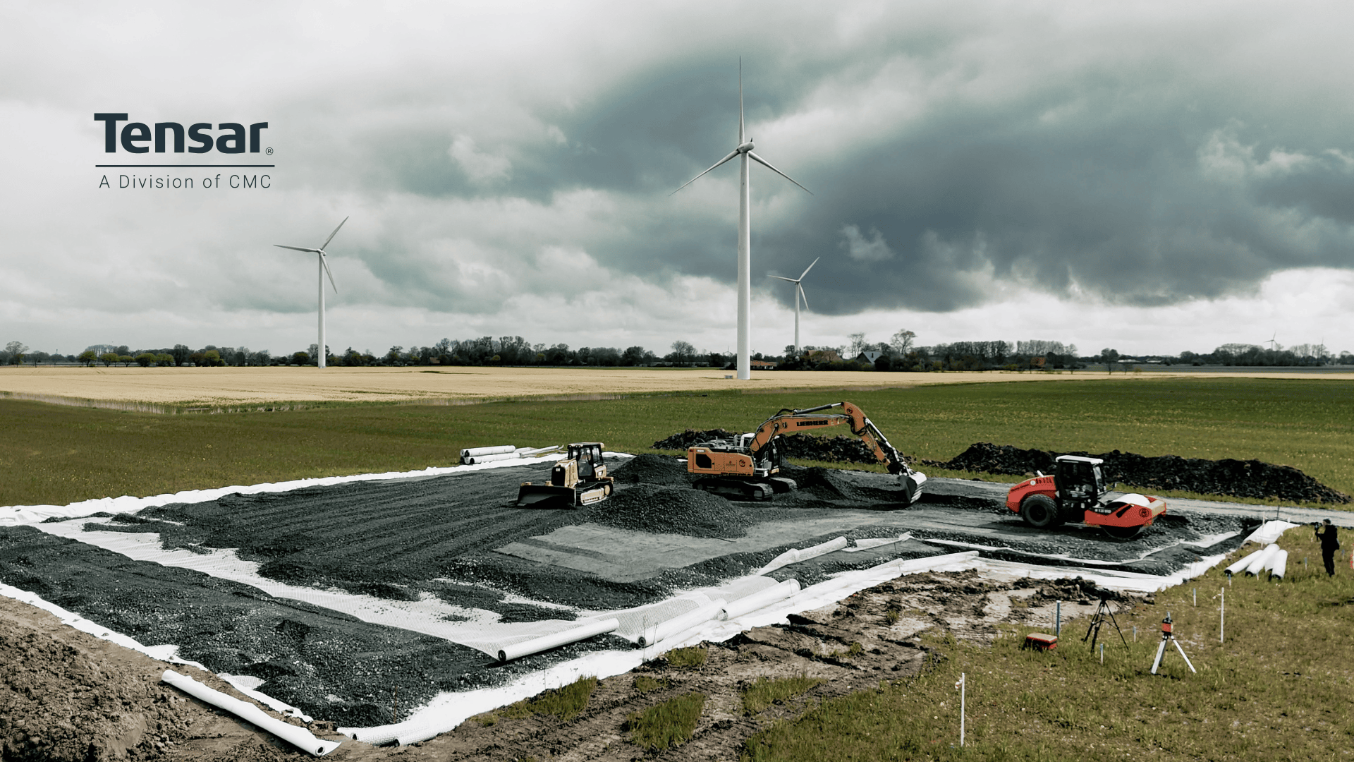 Heavy construction equipment—bulldozer, excavator, and compactor—work on a gravel foundation in a rural field. Three wind turbines and dramatic clouds are visible in the background. Tensar, A Division of CMC is on the left.