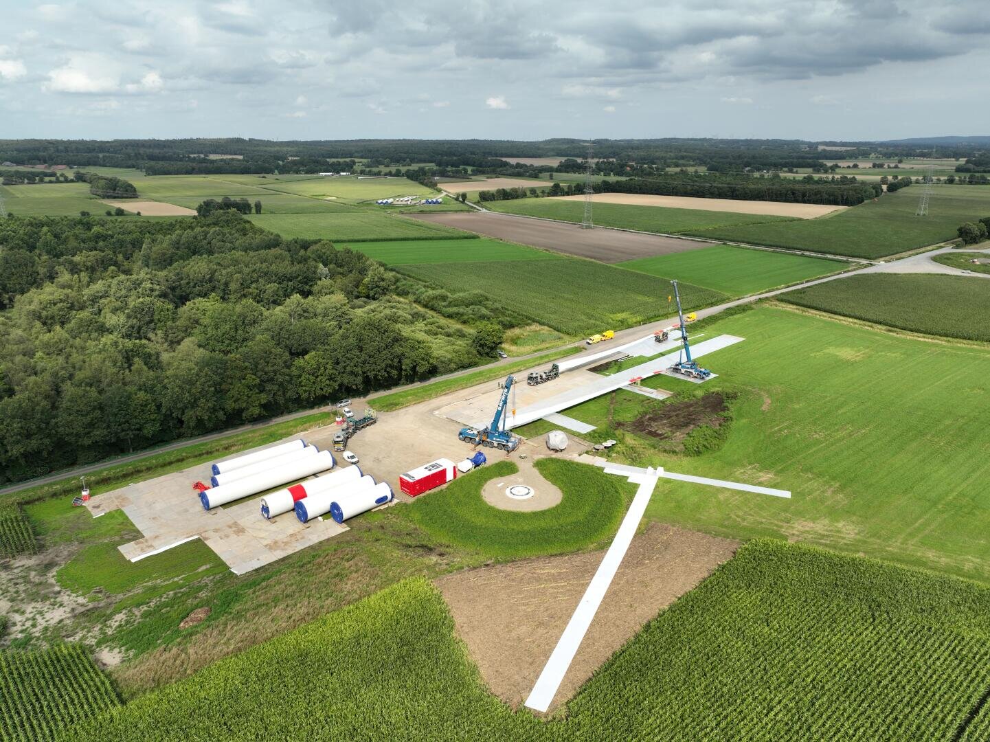 Luftaufnahme einer Windturbinen-Baustelle in einer ländlichen Gegend, mit großen Turbinenteilen, Kränen und Fahrzeugen, die auf dem Gras und dem Beton verstreut sind. Ackerland und Bäume umgeben die Baustelle unter einem bewölkten Himmel.