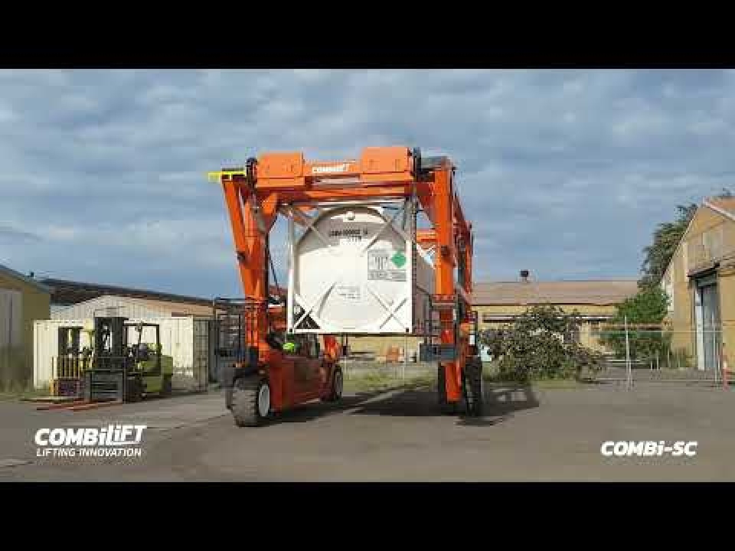 An orange Combilift straddle carrier lifts and transports a large white shipping container in an industrial yard, with warehouses and equipment visible in the background.