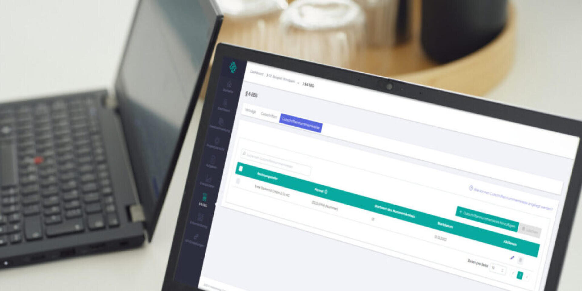 Ein Laptop, auf dem ein Software-Dashboard mit Tabellen und Navigationsmenüs angezeigt wird, steht auf einem Schreibtisch neben einem geschlossenen schwarzen Laptop, mit einem verschwommenen Tablett im Hintergrund.