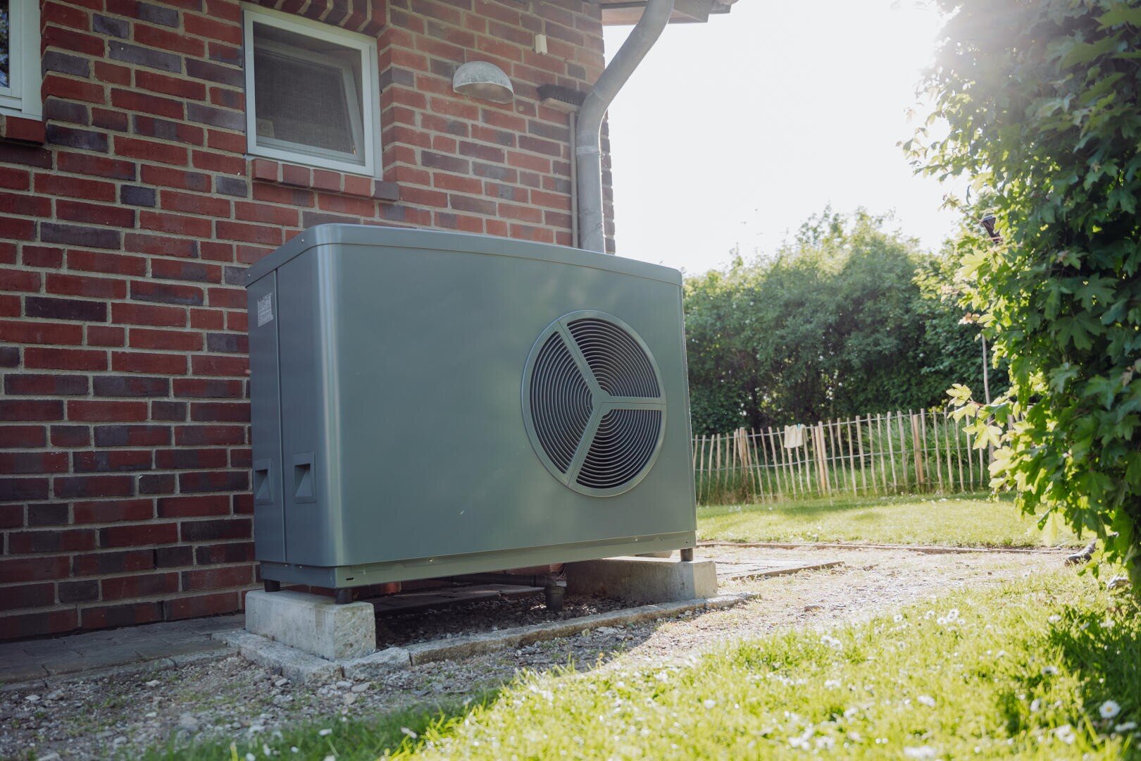An einem sonnigen Tag steht eine graue Wärmepumpeneinheit auf Betonblöcken neben einem Backsteinhaus, mit einem Fenster darüber und einem grasbewachsenen Hof mit Holzzaun in der Nähe.