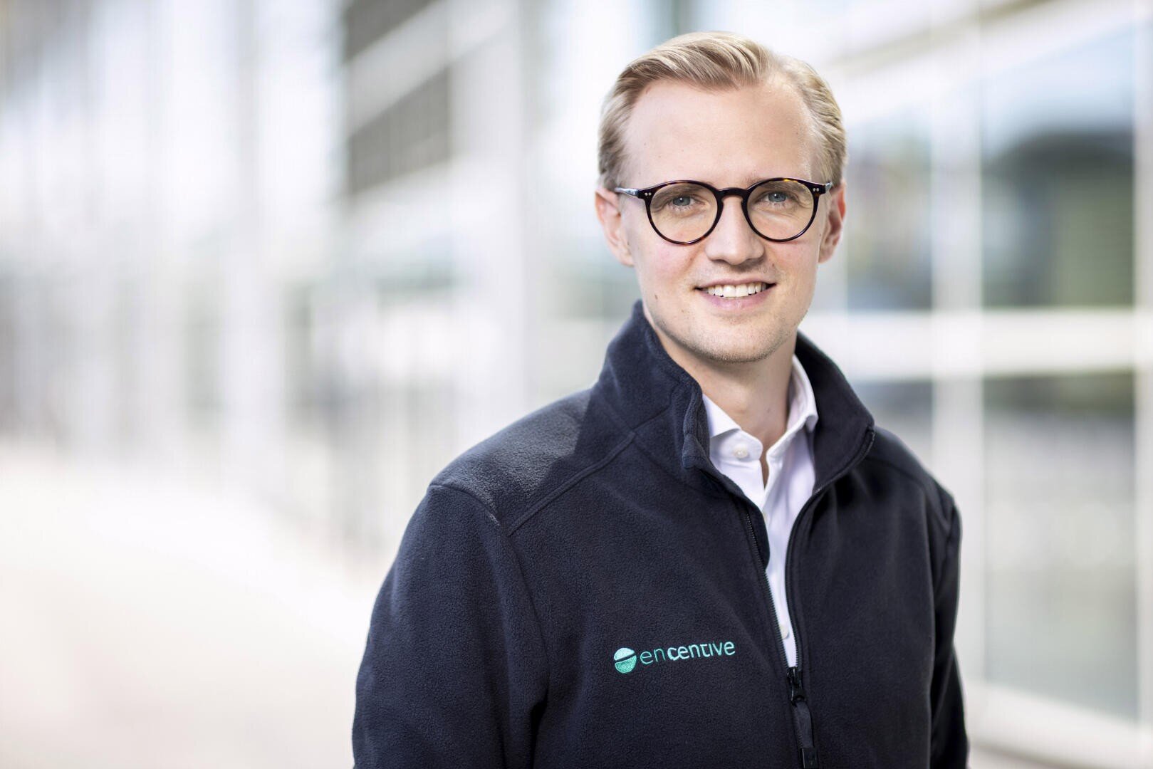A smiling man with blond hair and glasses wearing a dark jacket with the enCentive logo stands in front of a blurred modern glass building.
