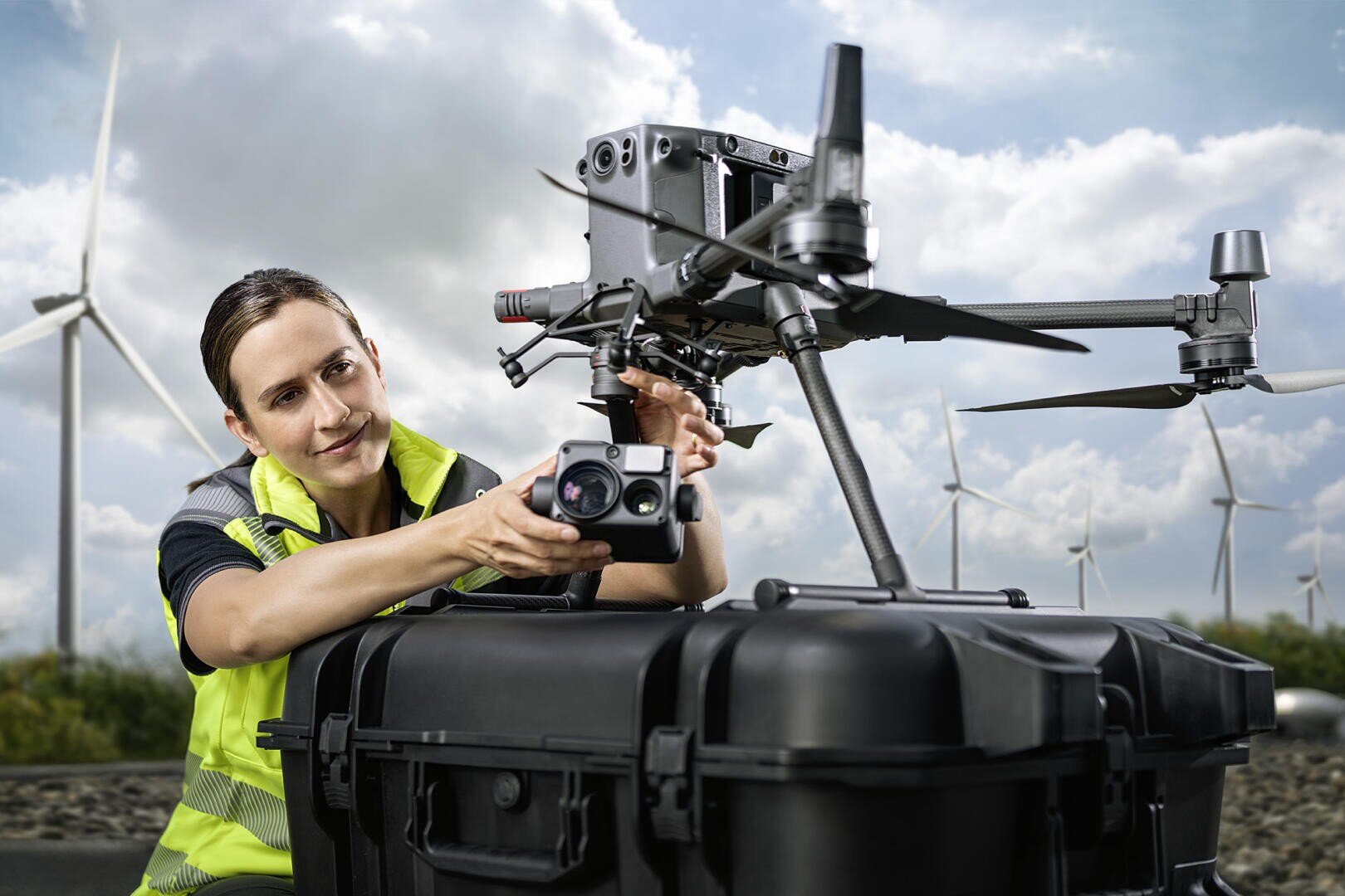 Eine Frau in einer Sicherheitsweste inspiziert eine große Drohne auf einem schwarzen Gehäuse. Im Hintergrund sind Windturbinen und ein bewölkter Himmel zu sehen, was auf erneuerbare Energien oder eine Inspektion hindeutet.
