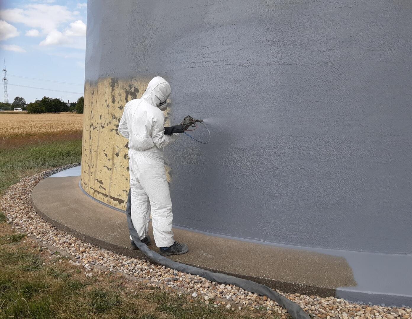 Eine Person in einem weißen Schutzanzug sprüht graue Farbe auf die Außenseite einer großen zylindrischen Struktur im Freien, mit Feldern und einem teilweise bewölkten Himmel im Hintergrund.