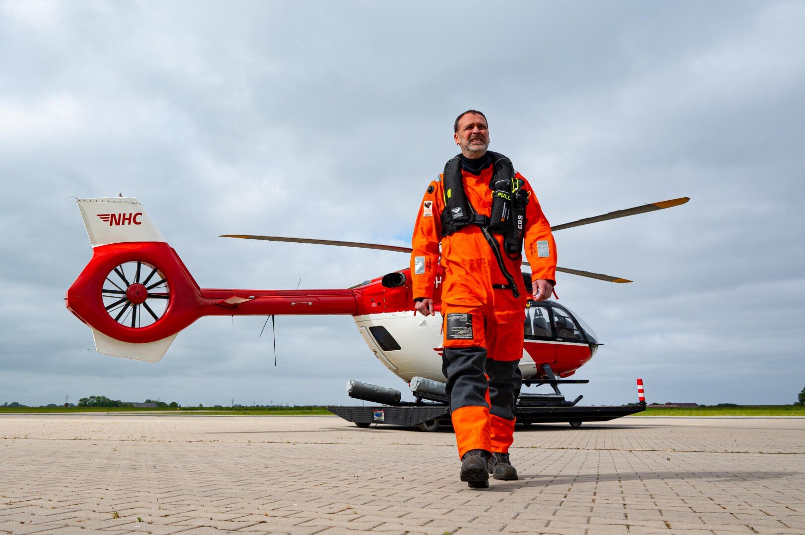 Ein Mann in einem orangefarbenen Fliegeranzug und einer Schwimmweste steht auf einer gepflasterten Fläche vor einem rot-weißen Rettungshubschrauber unter einem bewölkten Himmel.