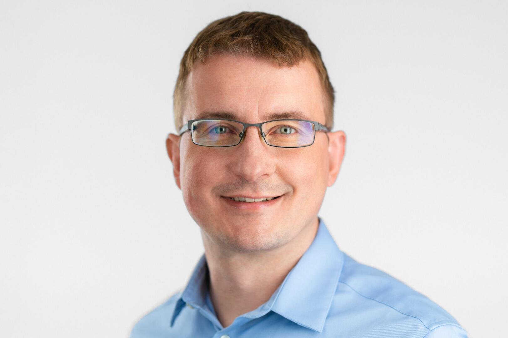 A man with short brown hair and glasses, wearing a light blue collared shirt, smiles at the camera against a plain white background.