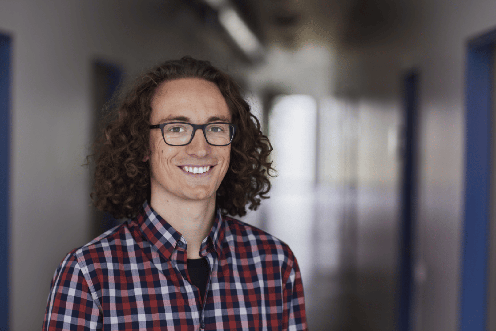Eine Person mit lockigem braunem Haar und Brille lächelt, während sie ein rot und marineblau kariertes Hemd trägt und in einem Flur mit verschwommenen blauen Türen und hellem Licht im Hintergrund steht.