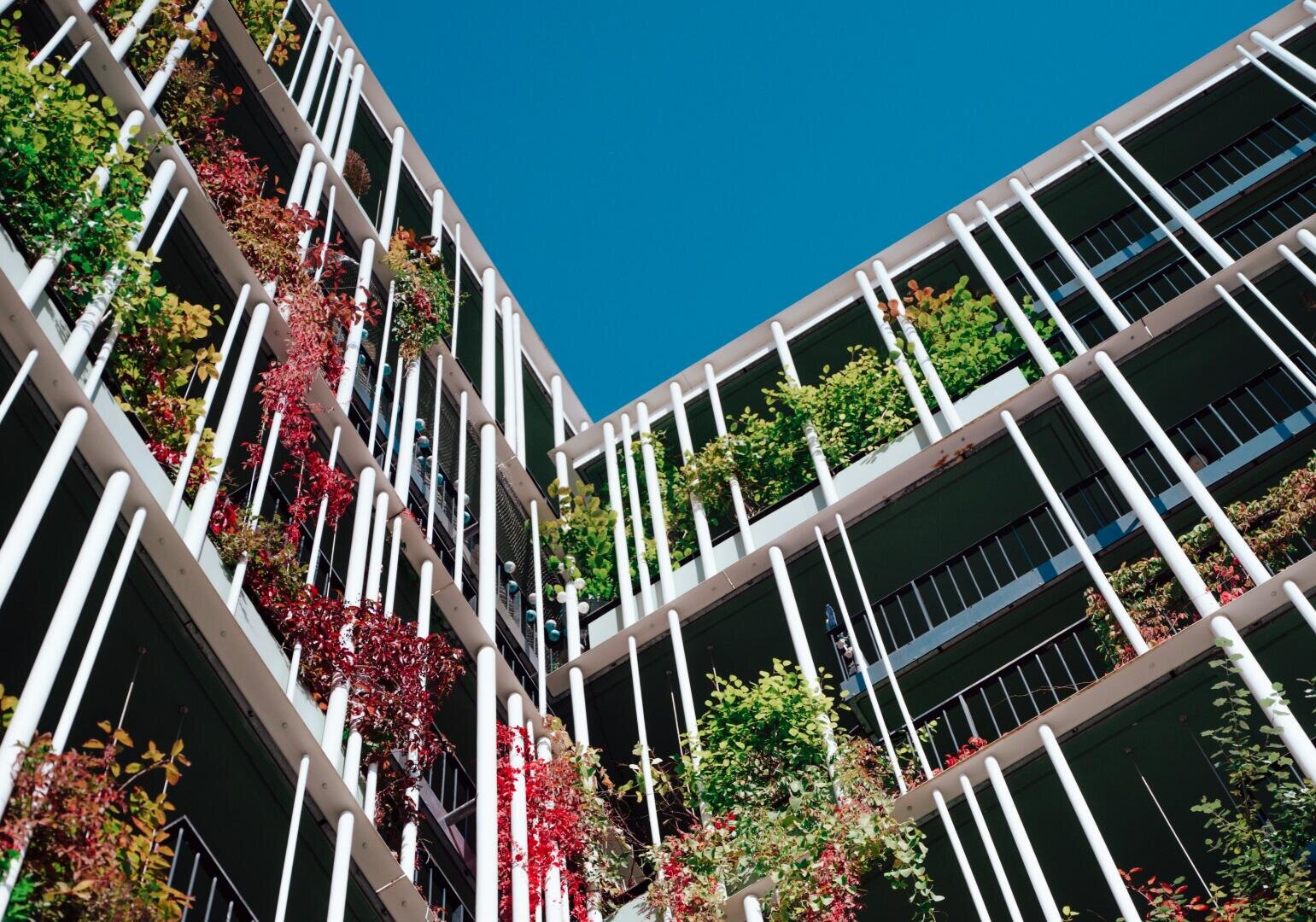 Ein modernes Wohnhaus mit grün und rot bepflanzten Balkonen, weißen vertikalen Säulen und einem klaren blauen Himmel darüber. Das Foto ist aus einem niedrigen Winkel aufgenommen und zeigt das Gebäude von oben.