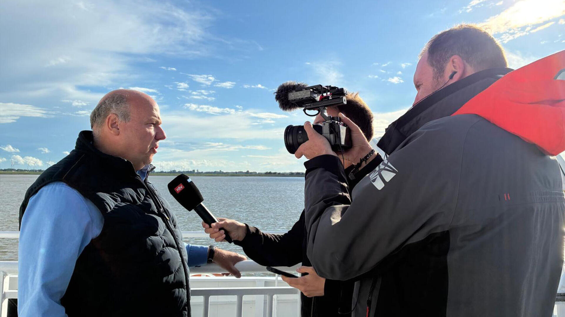 Ein Fernsehjournalist interviewt einen Mann auf einer Fähre, im Hintergrund ist ein Fluss zu sehen.