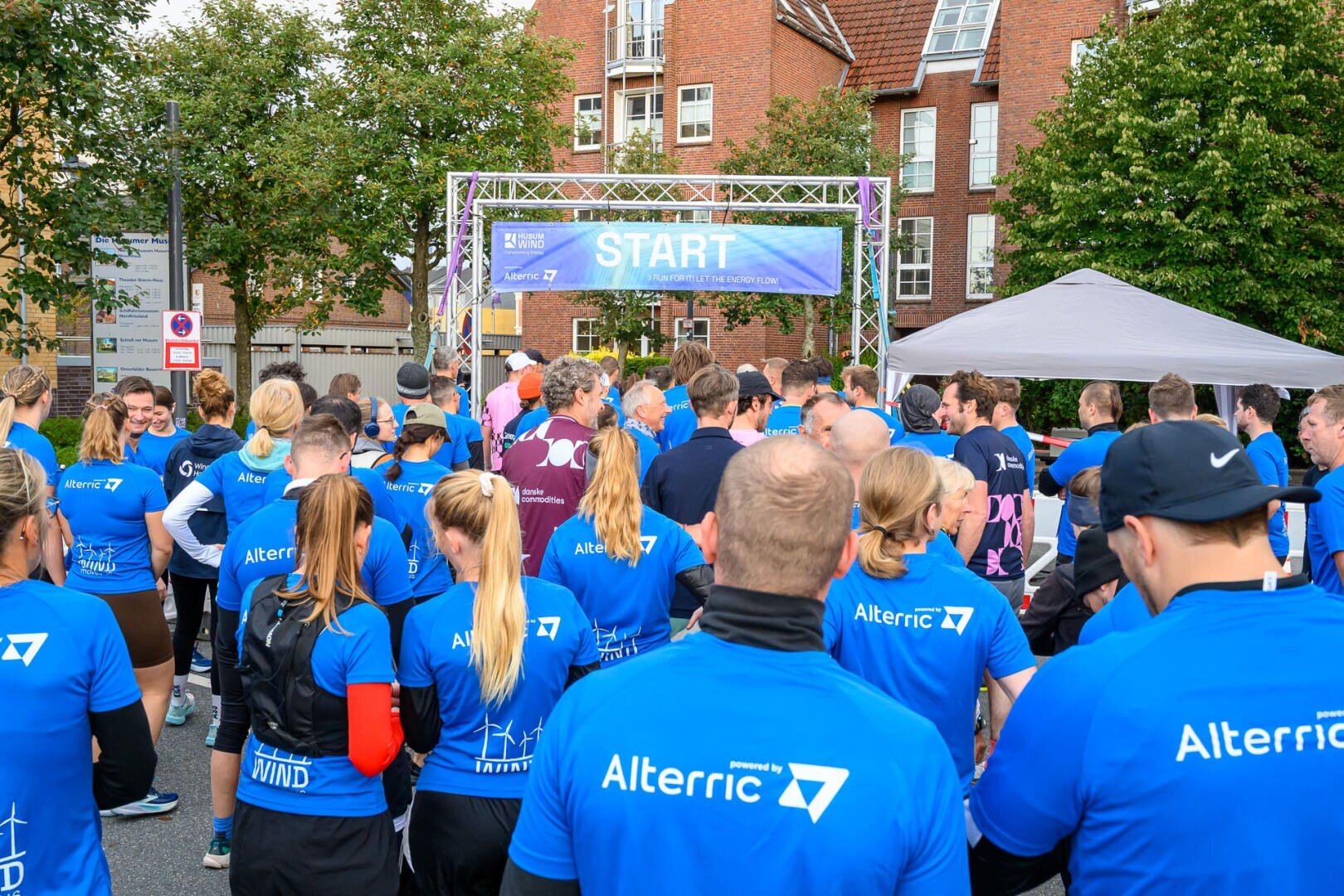 A crowd of people in matching blue shirts with the word "Alterric" on the back gather at the starting line for a race, with trees and brick buildings in the background. A banner above reads "START".